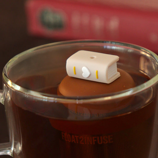 beige book with a white heart on the spine tea infuser for loose leaf tea with a fine mesh stainless steel tea strainer that reads FLOAT2INFUSEon a brown base  floating in a glass tea mug with books in the background