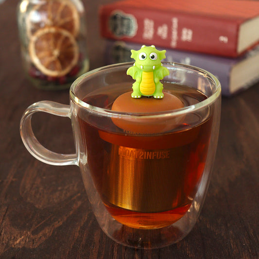 bright green dragon tea infuser for loose leaf tea  with a fine mesh stainless steel tea strainer that reads FLOAT2INFUSE on a brown base floating in a glass tea mug with a loose leaf tea and books in the background
