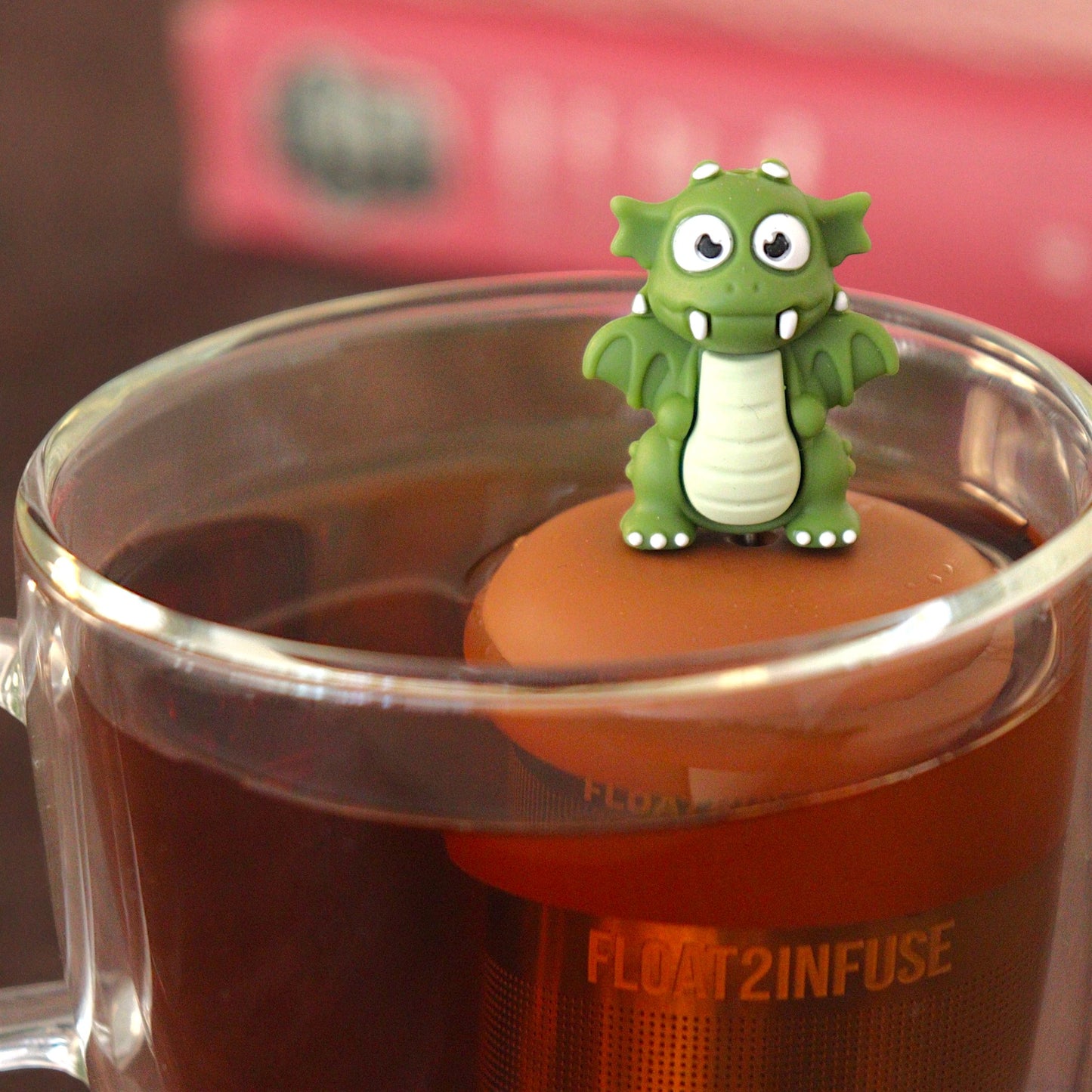 dark green dragon tea infuser for loose leaf tea  with a fine mesh stainless steel tea strainer that reads FLOAT2INFUSE on a brown base floating in a glass tea mug with  books in the background