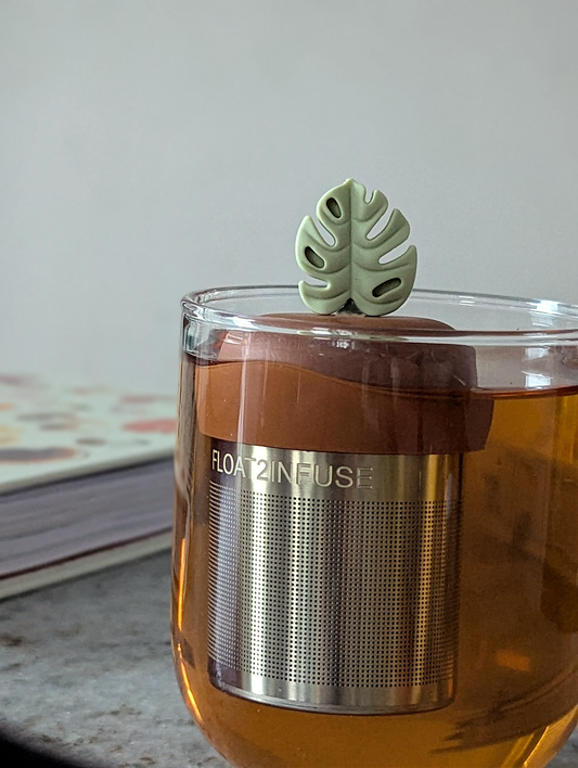 earth green monstera leaf tea infuser for loose leaf tea  with a fine mesh stainless steel tea steeper that reads FLOAT2INFUSE on a brown base floating in a glass tea mug with books in the background