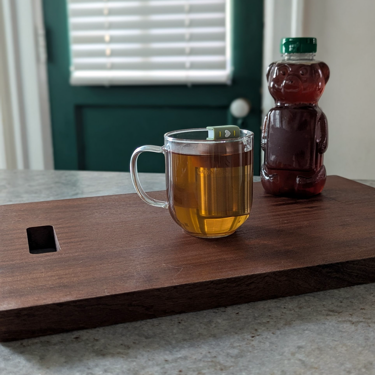 green book with a white heart on the spine tea infuser for loose leaf tea with fine mesh stainless steel tea strainer that reads FLOAT2INFUSE on a brown base floating in a glass tea mug with a honeybearin the background