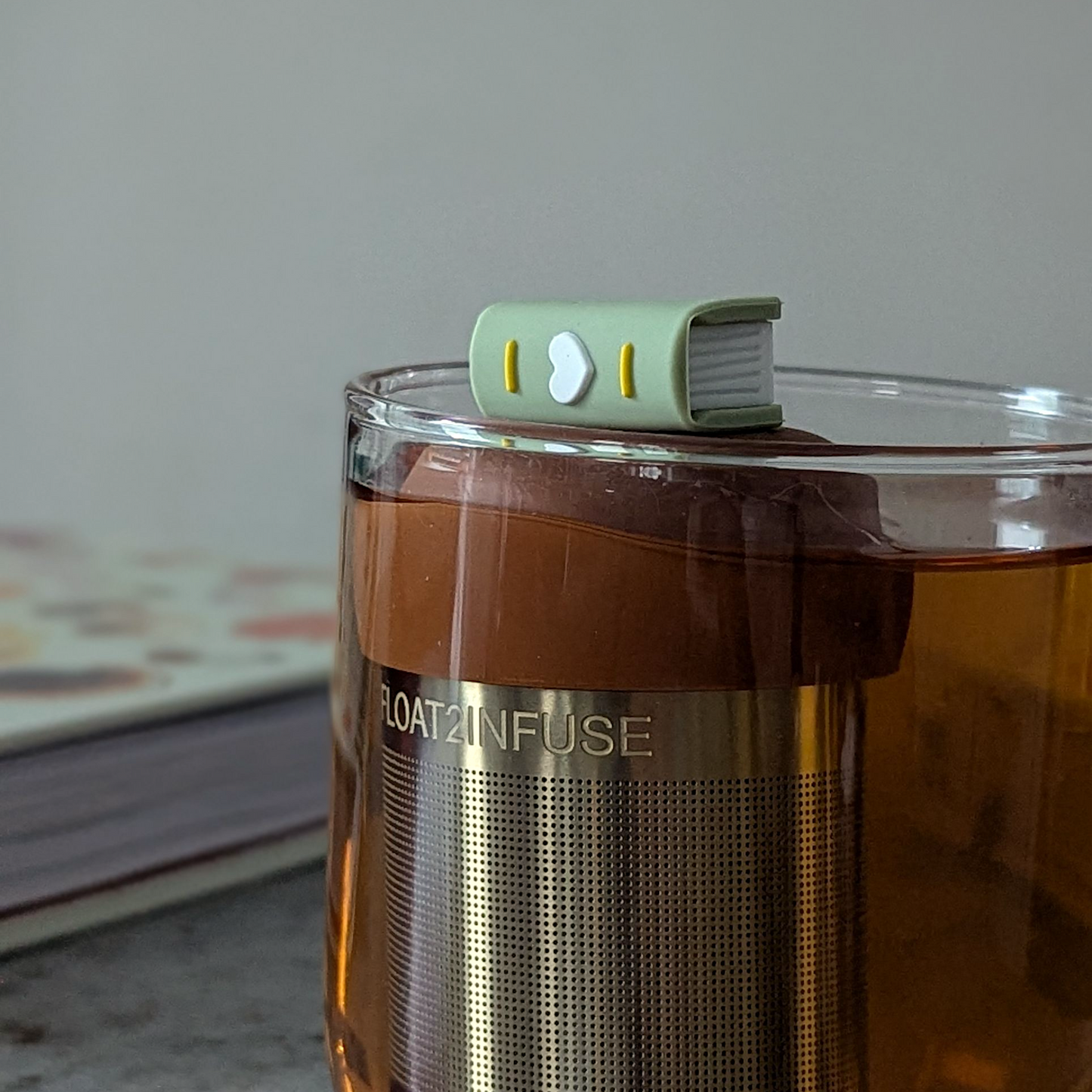 green book with a white heart on the spine tea infuser for loose leaf tea with fine mesh stainless steel tea strainer that reads FLOAT2INFUSE on a brown base floating in a glass tea mug with a book in the background