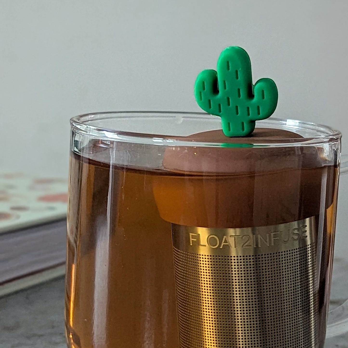 green cactus tea infuser for loose leaf tea  with a fine mesh stainless steel tea steeper that reads FLOAT2INFUSE on a brown base floating in a glass tea mug with books in the background