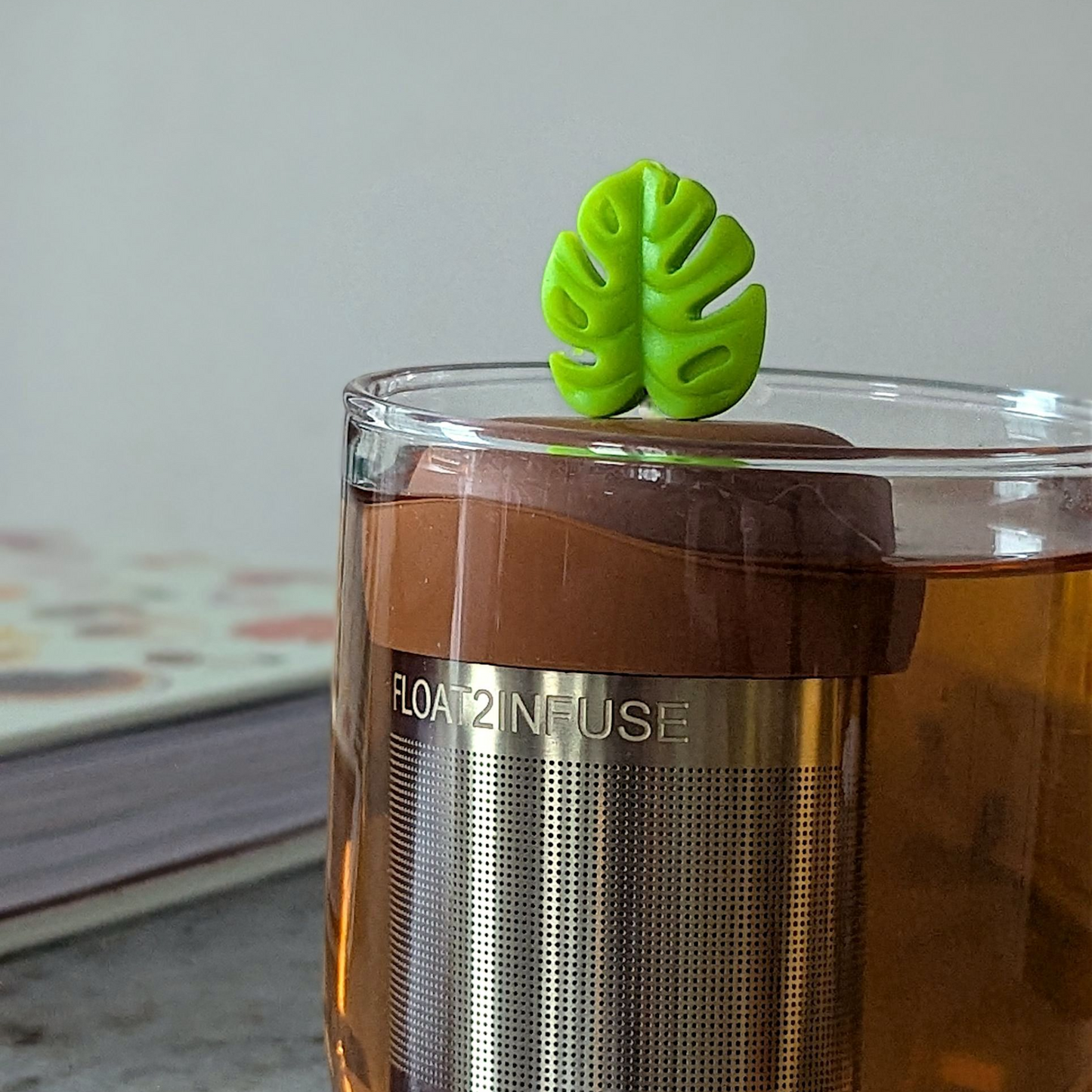 lime green monstera leaf tea infuser for loose leaf tea  with a fine mesh stainless steel tea steeper that reads FLOAT2INFUSE on a brown base floating in a glass tea mug with books in the background