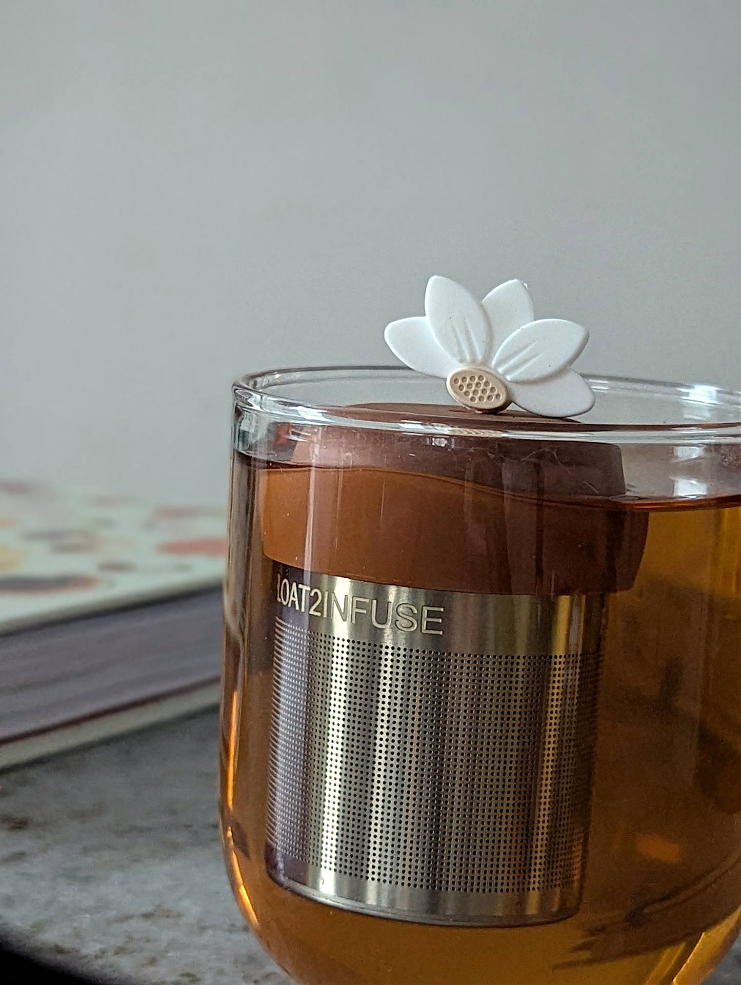 white lotus flower tea infuser for loose leaf tea with fine mesh stainless steel tea strainer that reads FLOAT2INFUSE on a brown base floating in a glass tea mug with a book in the background
