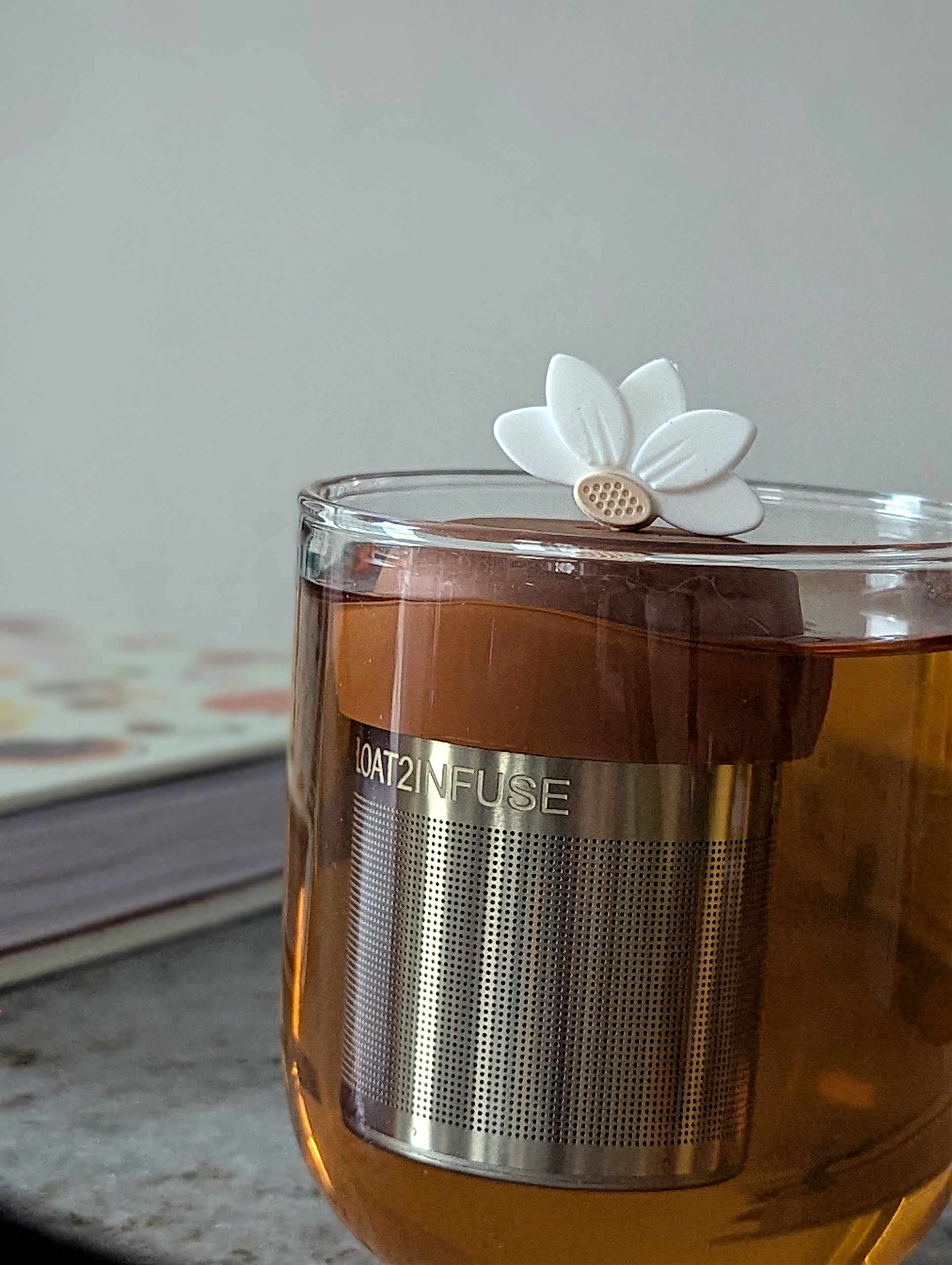 white lotus flower tea infuser for loose leaf tea with fine mesh stainless steel tea strainer that reads FLOAT2INFUSE on a brown base floating in a glass tea mug with a book in the background