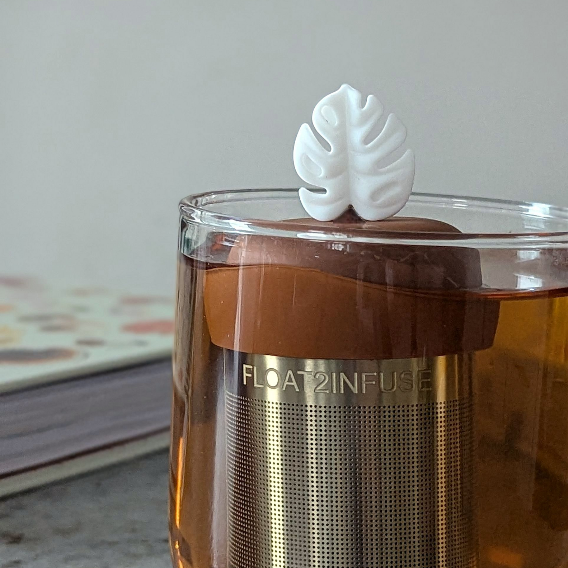 white monstera leaf tea infuser for loose leaf tea  with a fine mesh stainless steel tea steeper that reads FLOAT2INFUSE on a brown base floating in a glass tea mug with books in the background