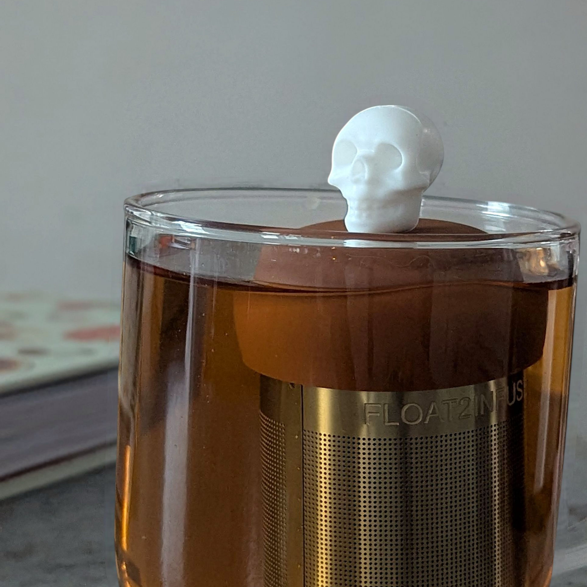 white skull tea infuser for loose leaf tea with fine mesh stainless steel tea strainer that reads FLOAT2INFUSE on a brown base floating in a glass tea mug with a book in the background