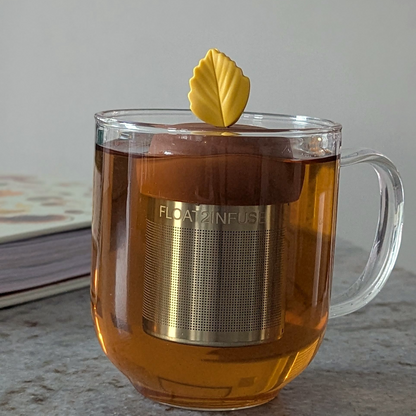 yellow fall leaf tea infuser for loose leaf tea with fine mesh stainless steel tea strainer that reads FLOAT2INFUSE on a brown base floating in a glass tea mug with a book in the background