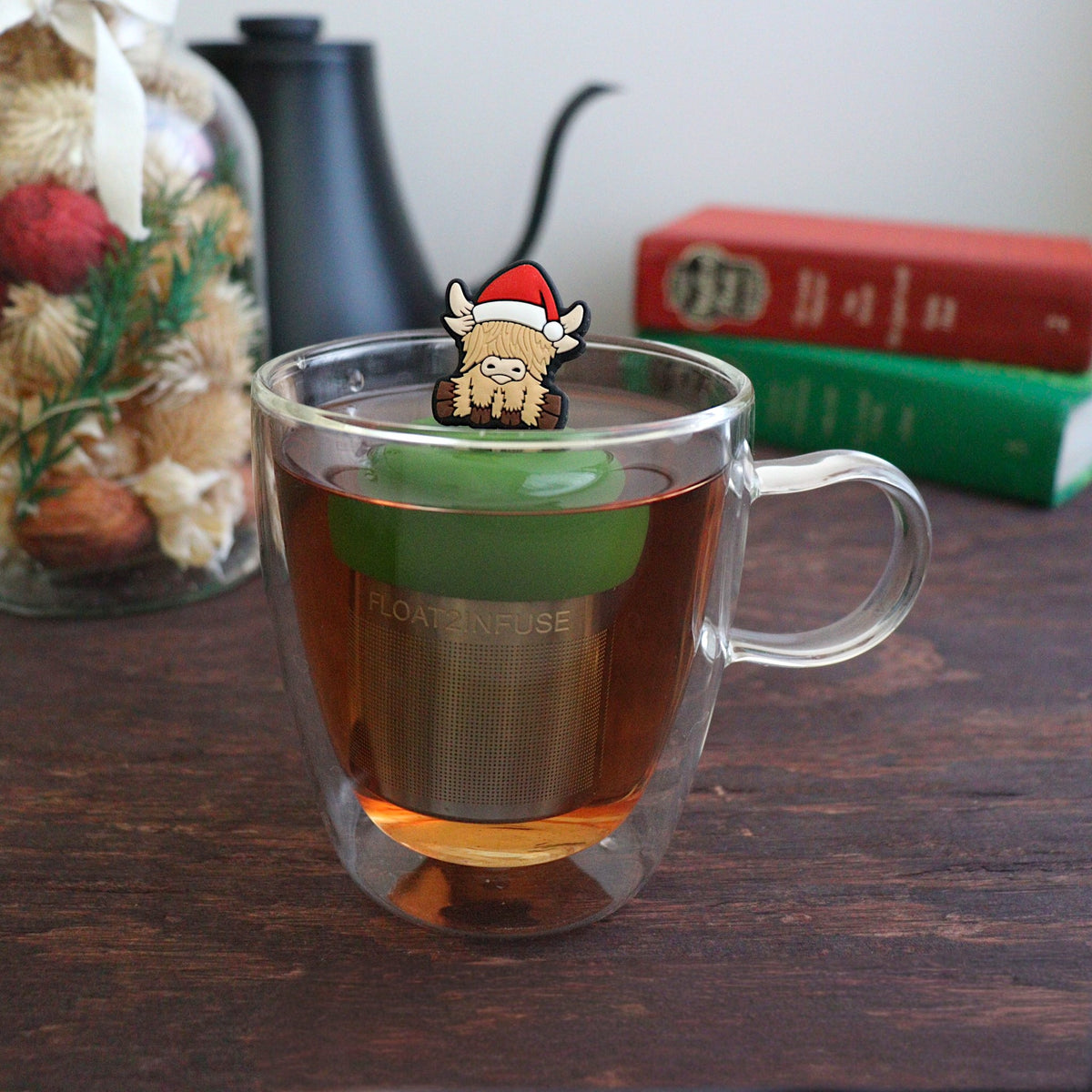 blonde highland cow animal wearing a santa hat tea infuser for loose leaf tea with a fine mesh stainless steel tea strainer that reads FLOAT2INFUSE on a dark green base floating in a glass tea mug with books and Christmas decorations in the background