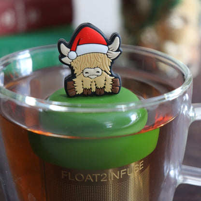blonde highland cow animal wearing a santa hat tea infuser for loose leaf tea with a fine mesh stainless steel tea strainer that reads FLOAT2INFUSE on a dark green base floating in a glass tea mug with books and Christmas decorations in the background