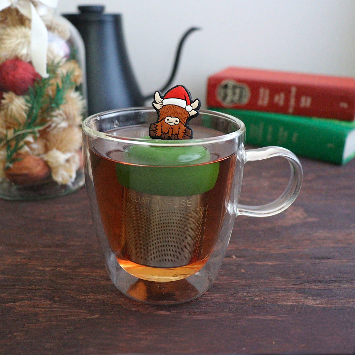 Brown highland cow wearing a santa hat a  tea infuser for loose leaf tea with a fine mesh stainless steel tea strainer  that reads FLOAT2INFUSE on a dark green base with books, a tea kettle, and christmas decorations in the background