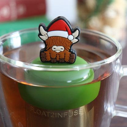 Brown highland cow wearing a santa hat a  tea infuser for loose leaf tea with a fine mesh stainless steel tea strainer  that reads FLOAT2INFUSE on a dark green base with books, a tea kettle, and christmas decorations in the background