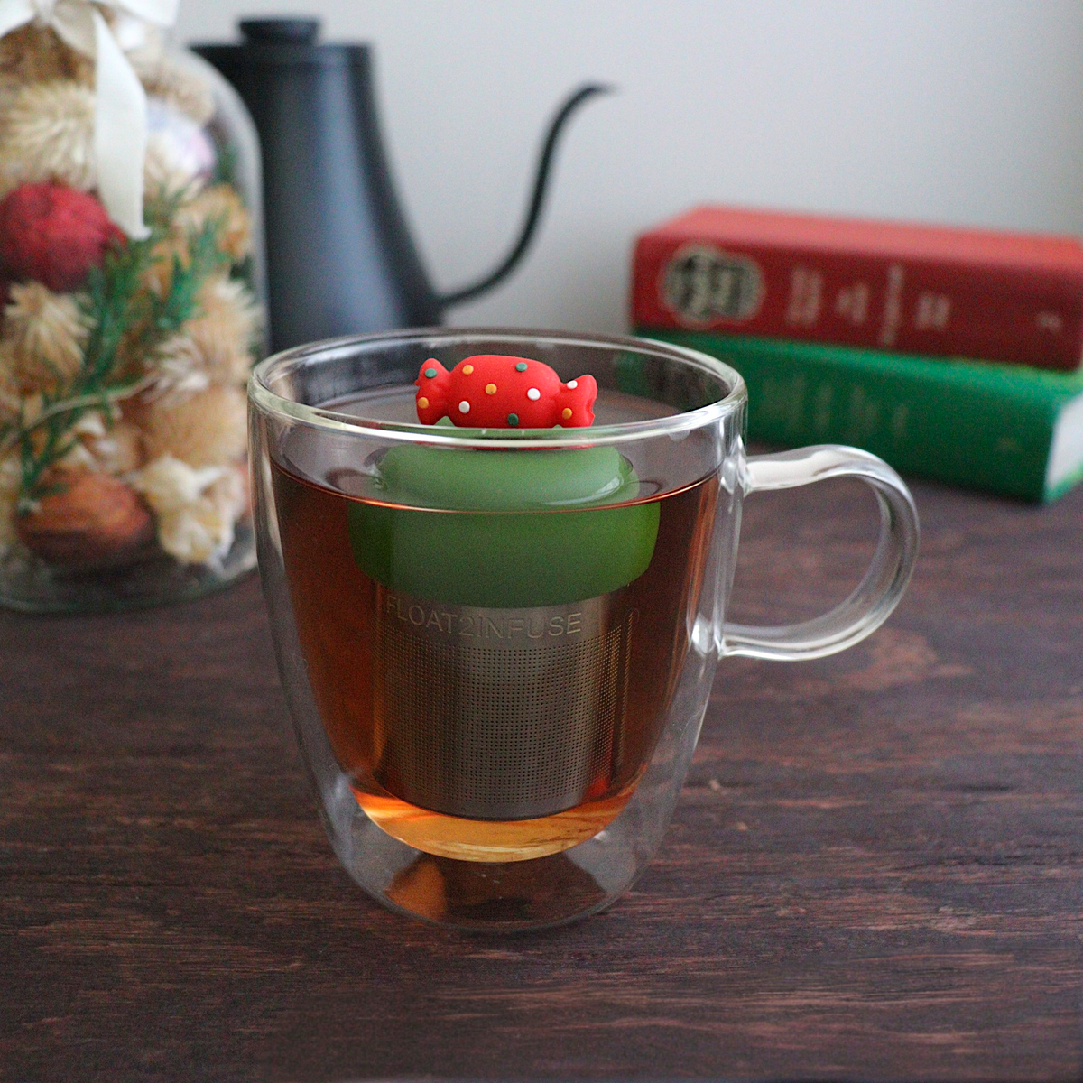  Red holiday candy with green, yellow, and white polka dots for loose leaf tea with a fine mesh stainless steel tea strainer that reads FLOAT2INFUSE on a dark green base with books, a tea kettle, and christmas decorations in the background