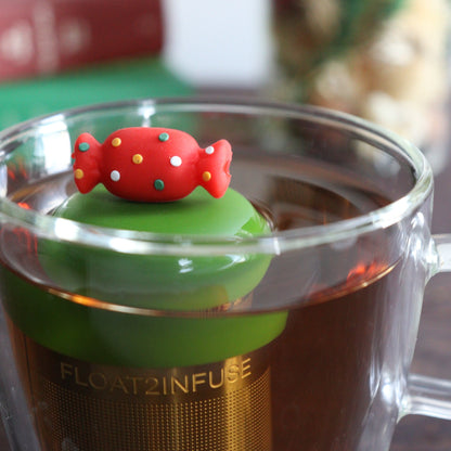  Red holiday candy with green, yellow, and white polka dots for loose leaf tea with a fine mesh stainless steel tea strainer that reads FLOAT2INFUSE on a dark green base with books, a tea kettle, and christmas decorations in the background