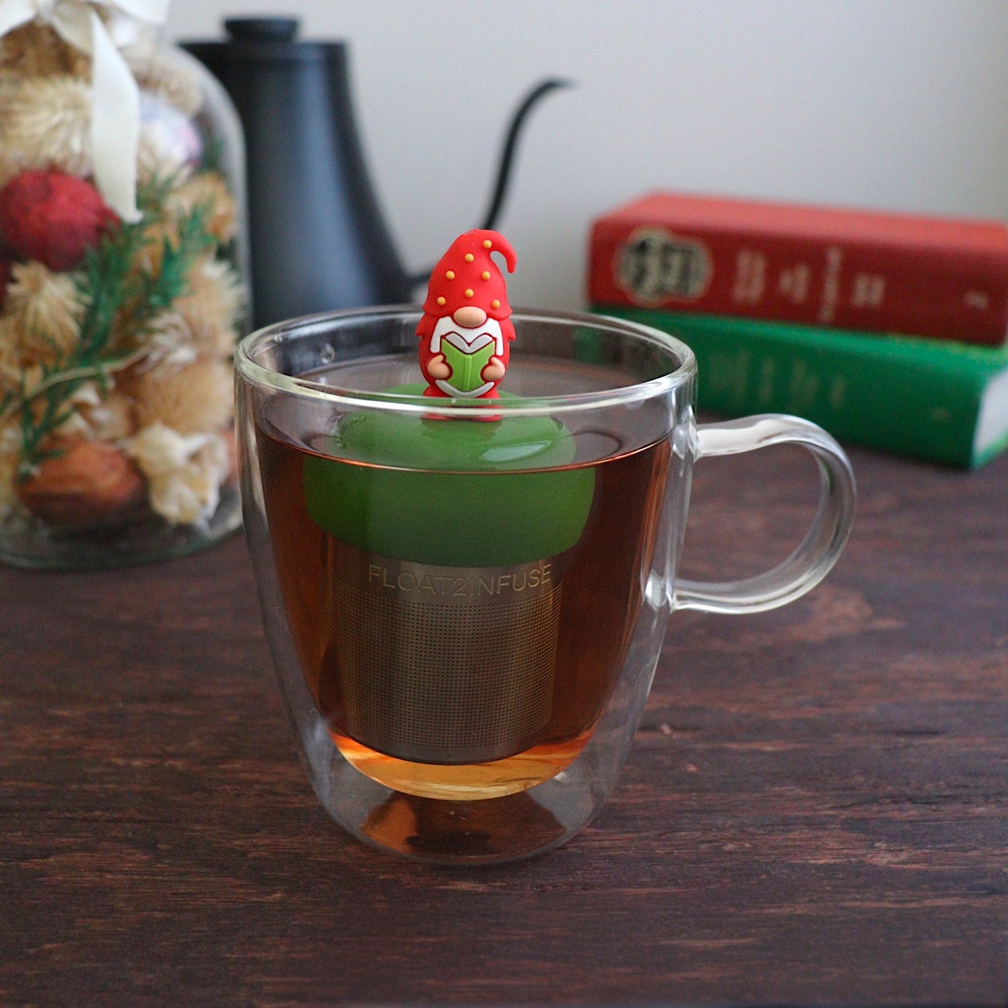 red holiday candy with green, yellow, and white polka dots tea infuser for loose leaf tea with a fine mesh stainless steel tea strainer that reads FLOAT2INFUSE on a dark green base with books, a tea kettle, and christmas decorations in the background