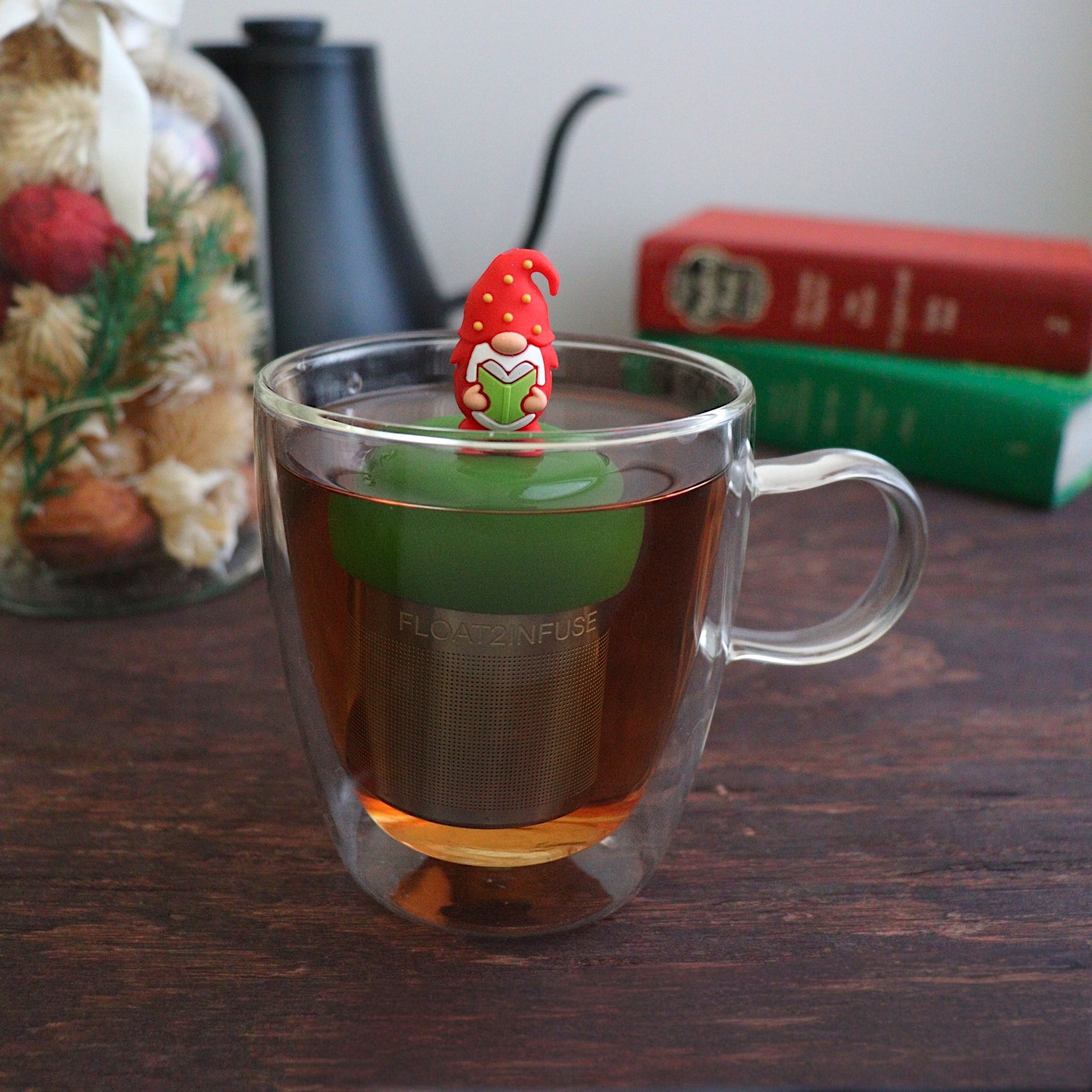 red holiday candy with green, yellow, and white polka dots tea infuser for loose leaf tea with a fine mesh stainless steel tea strainer that reads FLOAT2INFUSE on a dark green base with books, a tea kettle, and christmas decorations in the background