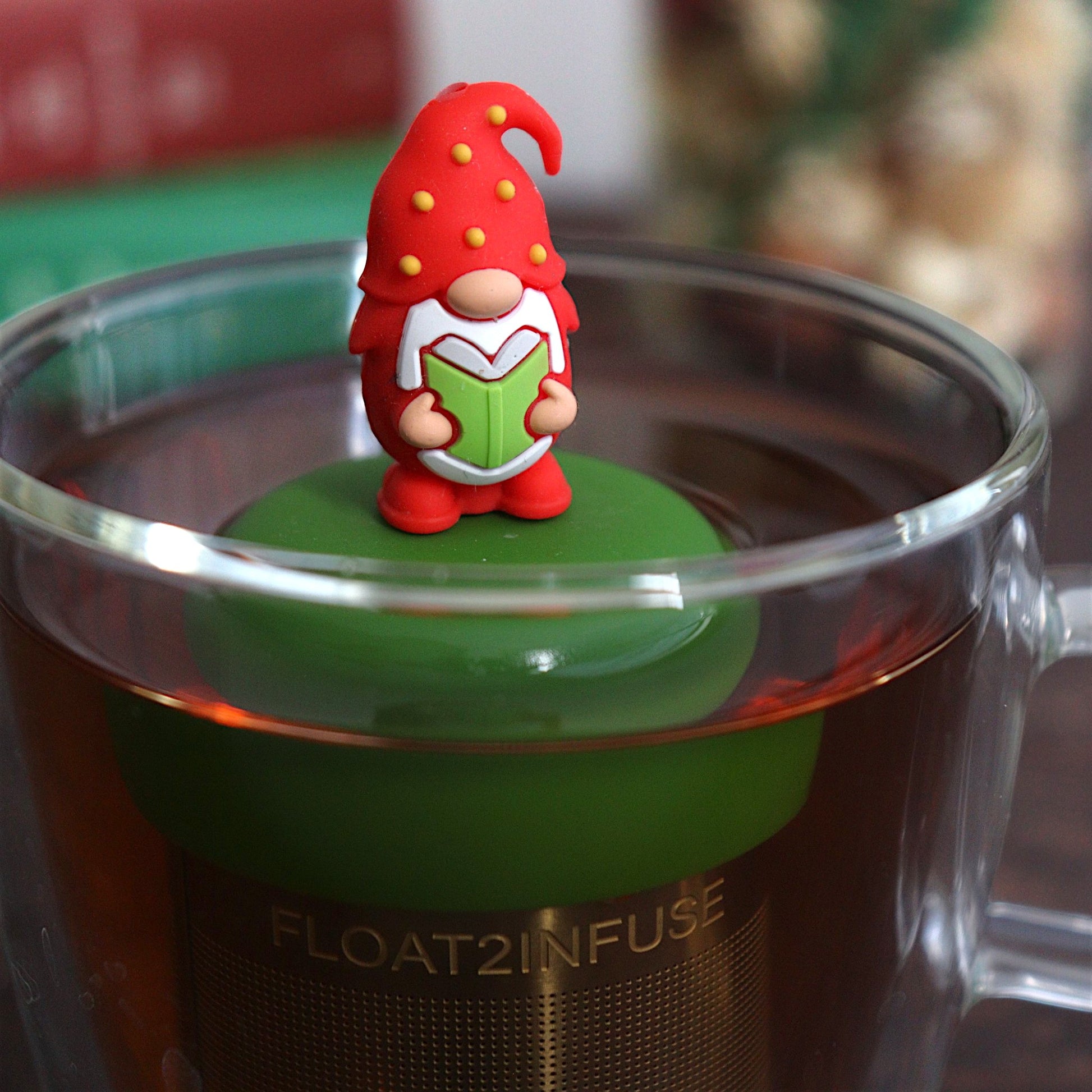 red holiday candy with green, yellow, and white polka dots tea infuser for loose leaf tea with a fine mesh stainless steel tea strainer that reads FLOAT2INFUSE on a dark green base with books, a tea kettle, and christmas decorations in the background