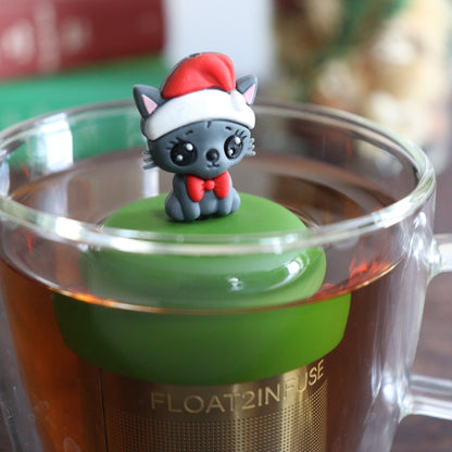 happy grey cat wearing a santa hat and red bowtie tea infuser for loose leaf tea with a fine mesh stainless steel tea strainer that reads FLOAT2INFUSE on a dark green base with books, a tea kettle, and christmas decorations in the background