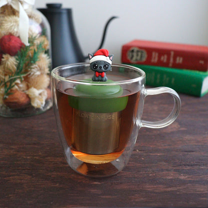 happy grey cat wearing a santa hat and red bowtie tea infuser for loose leaf tea with a fine mesh stainless steel tea strainer that reads FLOAT2INFUSE on a green base with books, a tea kettle, and christmas decorations in the background