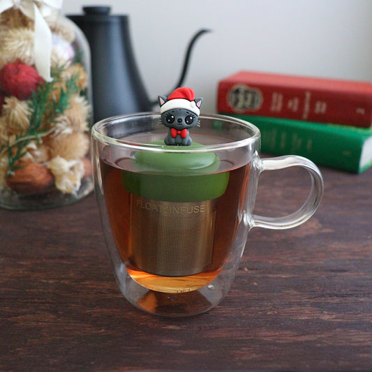 happy grey cat wearing a santa hat and red bowtie tea infuser for loose leaf tea with a fine mesh stainless steel tea strainer that reads FLOAT2INFUSE on a green base with books, a tea kettle, and christmas decorations in the background