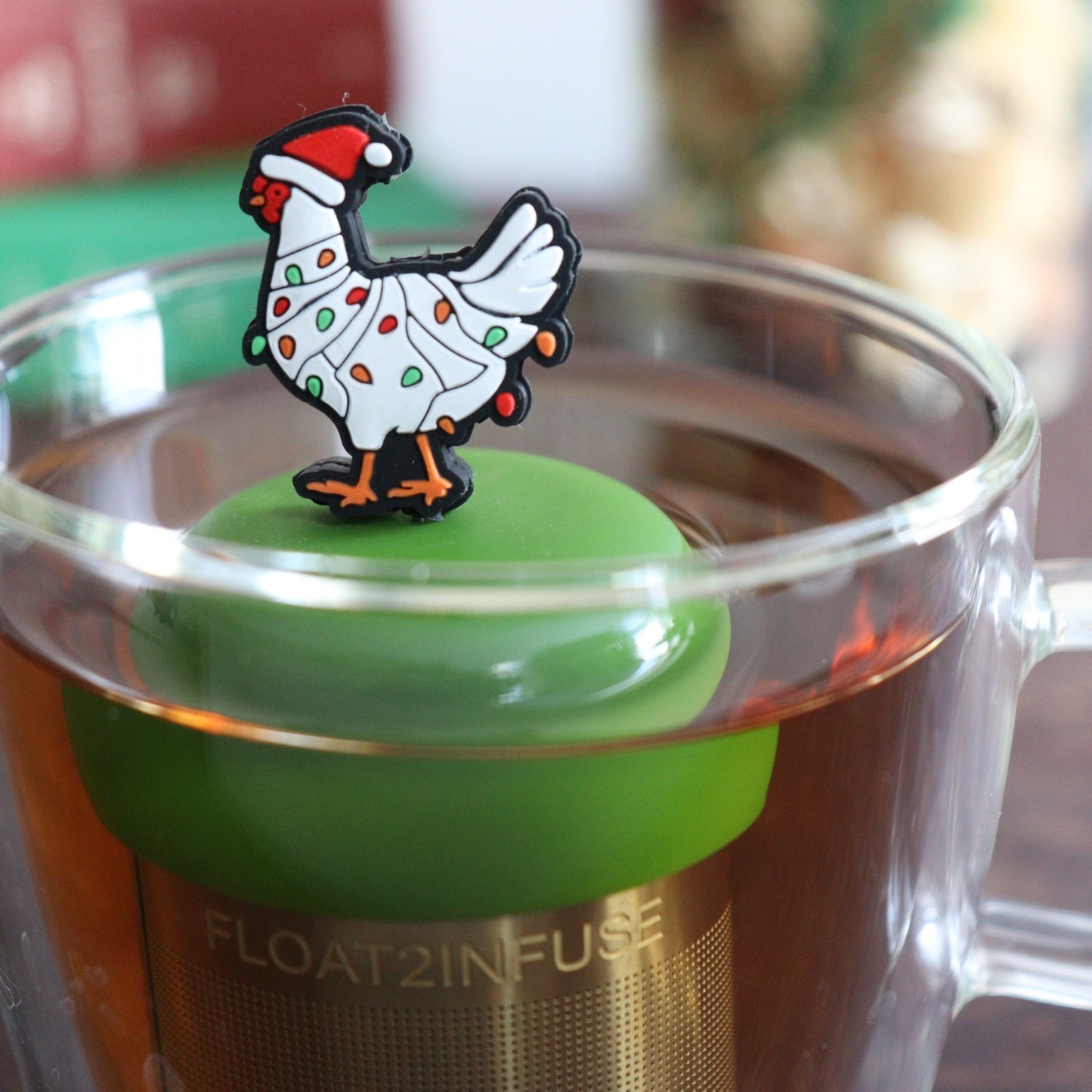 White chicken wearing a santa hat and wrapped in red and green christmas lights  tea infuser for loose leaf tea with a fine mesh stainless steel tea strainer  that reads FLOAT2INFUSE on a dark green base with books, a tea kettle, and christmas decorations in the background