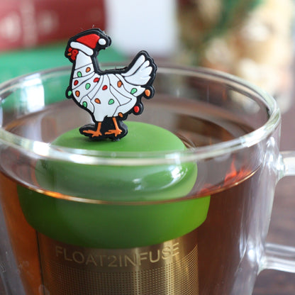 White chicken wearing a santa hat and wrapped in red and green christmas lights  tea infuser for loose leaf tea with a fine mesh stainless steel tea strainer  that reads FLOAT2INFUSE on a dark green base with books, a tea kettle, and christmas decorations in the background