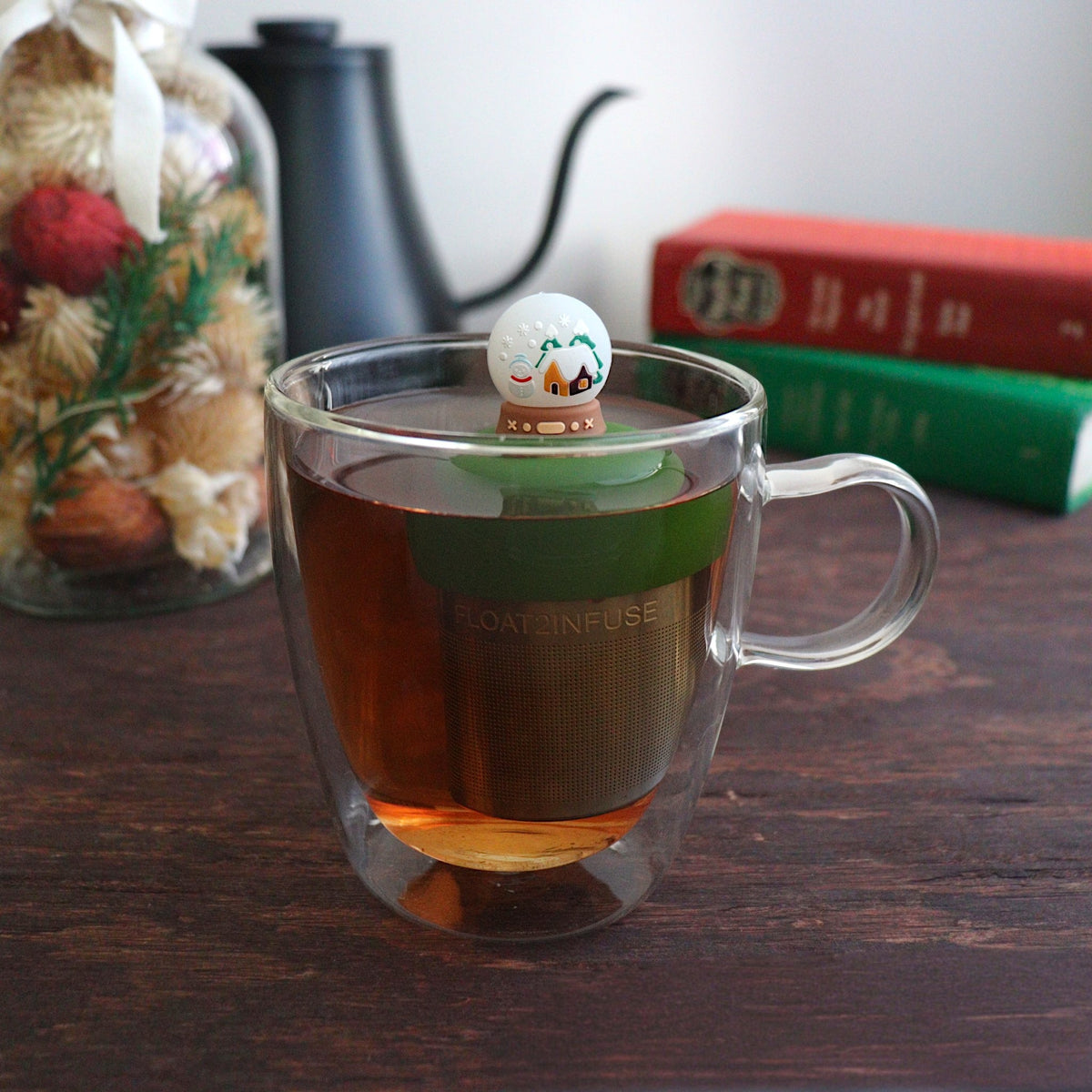 back view of a snowglobe with a snowman and a cabin inside for loose leaf tea with fine mesh stainless steel tea strainer that reads FLOAT2INFUSE on a dark green base floating in a glass tea mug with books and Christmas decorations in the background