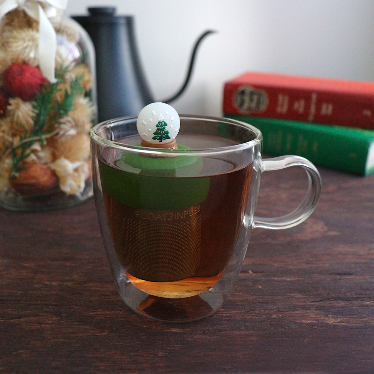front view of a snowglobe with a christmas tree inside for loose leaf tea with fine mesh stainless steel tea strainer that reads FLOAT2INFUSE on a dark green base floating in a glass tea mug with books and Christmas decorations in the background