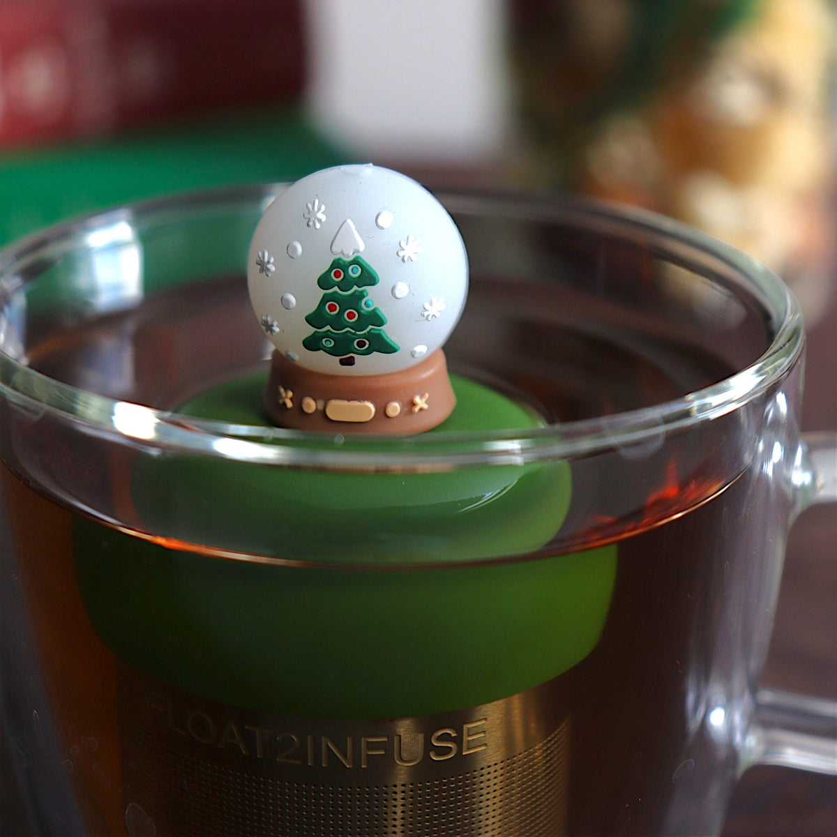 front view of a snowglobe with a christmas tree inside for loose leaf tea with fine mesh stainless steel tea strainer that reads FLOAT2INFUSE on a dark green base floating in a glass tea mug with books and Christmas decorations in the background