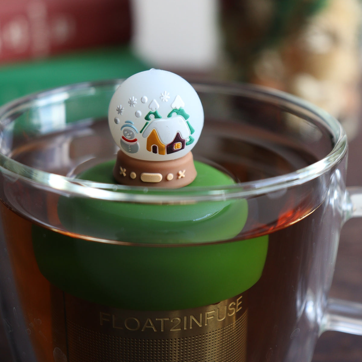 back view of a snowglobe with a snowman and a cabin inside for loose leaf tea with fine mesh stainless steel tea strainer that reads FLOAT2INFUSE on a dark green base floating in a glass tea mug with books and Christmas decorations in the background