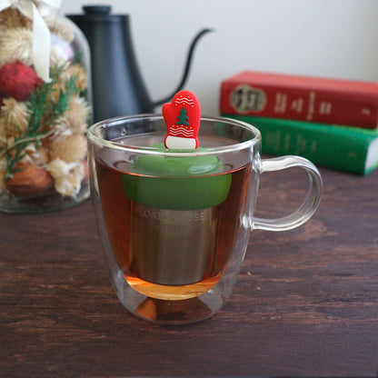 red mitten with green christmas tree and white accents tea infuser for loose leaf tea with a fine mesh stainless steel tea strainer that reads FLOAT2INFUSE on a dark green base  floating in a glass tea mug with books and Christmas decorations in the background