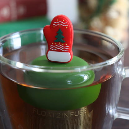 red mitten with green christmas tree and white accents tea infuser for loose leaf tea with a fine mesh stainless steel tea strainer that reads FLOAT2INFUSE on a dark green base  floating in a glass tea mug with books and Christmas decorations in the background