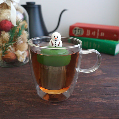 smiling fat snowman infuser for loose leaf tea with a fine mesh stainless steel tea strainer that reads FLOAT2INFUSE on a dark green base with a tea canister and books in the background