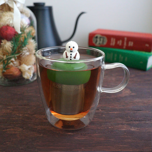 smiling fat snowman infuser for loose leaf tea with a fine mesh stainless steel tea strainer that reads FLOAT2INFUSE on a dark green base with a tea canister and books in the background
