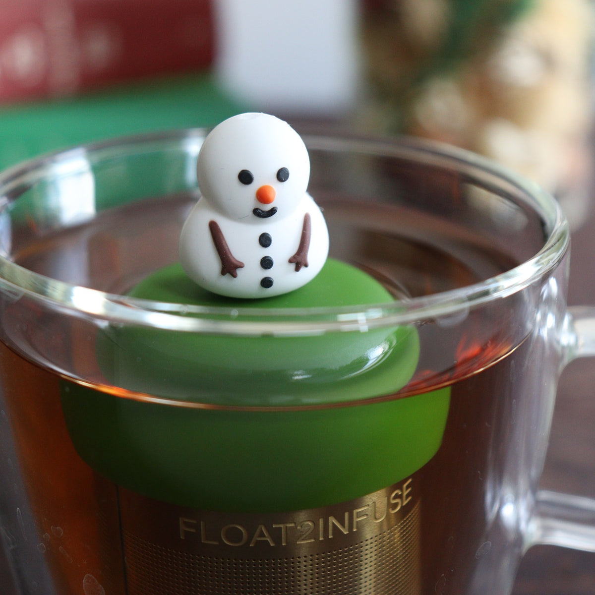 smiling fat snowman infuser for loose leaf tea with a fine mesh stainless steel tea strainer that reads FLOAT2INFUSE on a dark green base with a tea canister and books in the background