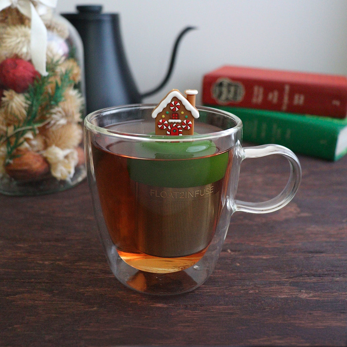Brown gingerbread house with peppermint candy windows and snow on the roof tea infuser for loose leaf tea with a fine mesh stainless steel tea strainer that reads FLOAT2INFUSE on a dark green base floating in a glass tea mug with books and Christmas decorations in the background