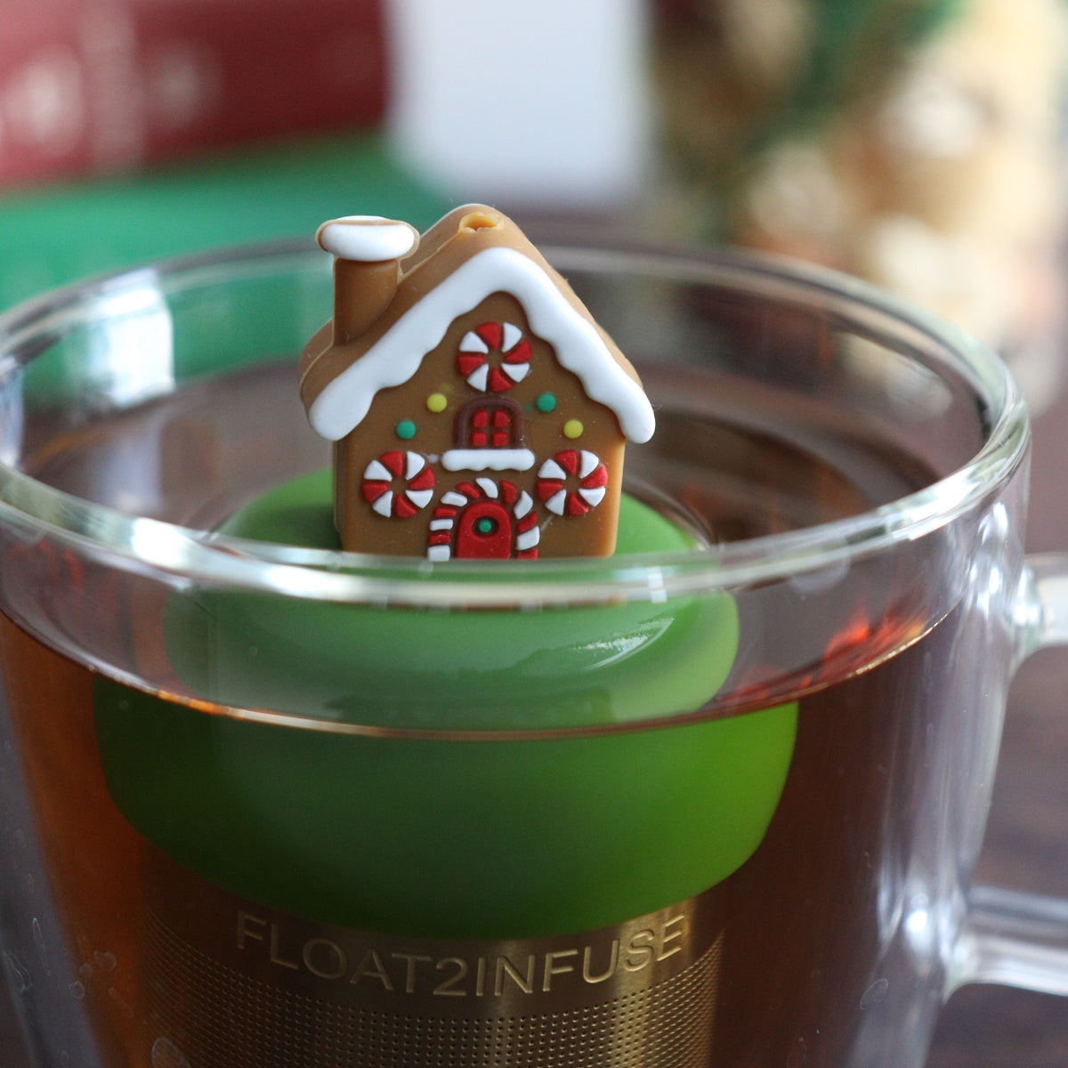 Brown gingerbread house with peppermint candy windows and snow on the roof tea infuser for loose leaf tea with a fine mesh stainless steel tea strainer that reads FLOAT2INFUSE on a dark green base floating in a glass tea mug with books and Christmas decorations in the background