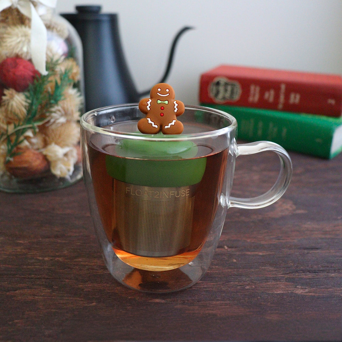 happy brown gingerbread man with green bowtie and red buttons tea infuser for loose leaf tea with a fine mesh stainless steel tea strainer that reads FLOAT2INFUSE on a dark green base with books, a tea kettle, and christmas decorations in the background