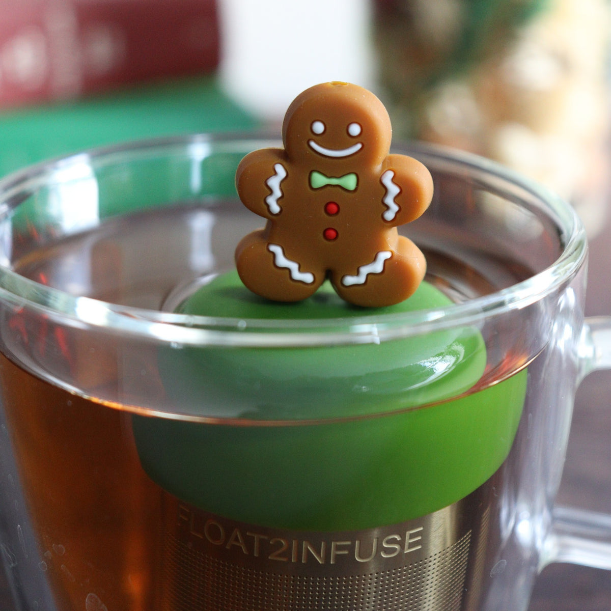 happy brown gingerbread man with green bowtie and red buttons tea infuser for loose leaf tea with a fine mesh stainless steel tea strainer that reads FLOAT2INFUSE on a dark green base with books, a tea kettle, and christmas decorations in the background