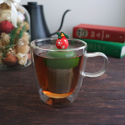 red christmas ornament with white snowflakes and green holly tea infuser for loose leaf tea with a fine mesh stainless steel tea strainer that reads FLOAT2INFUSE on a dark green base with books, a tea kettle, and christmas decorations in the background