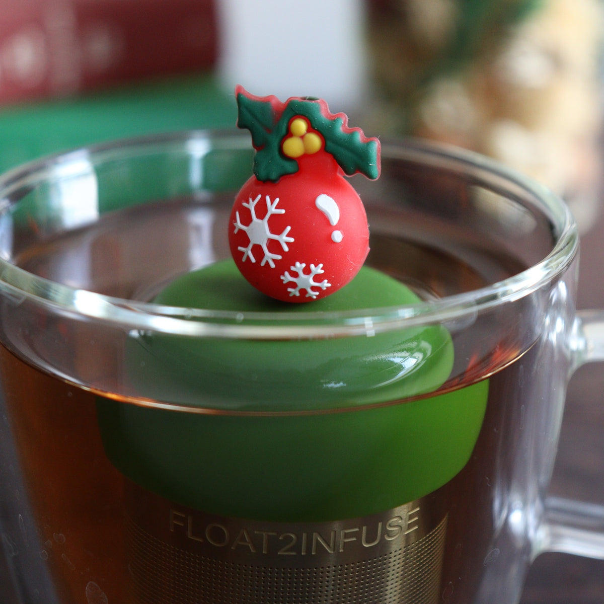 red christmas ornament with white snowflakes and green holly tea infuser for loose leaf tea with a fine mesh stainless steel tea strainer that reads FLOAT2INFUSE on a dark green base with books, a tea kettle, and christmas decorations in the background