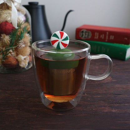 red, white, and green striped peppermint candy tea infuser for loose leaf tea with a fine mesh stainless steel tea strainer 
that reads FLOAT2INFUSE on a dark green base floating in a glass tea mug with books and Christmas decorations in the background