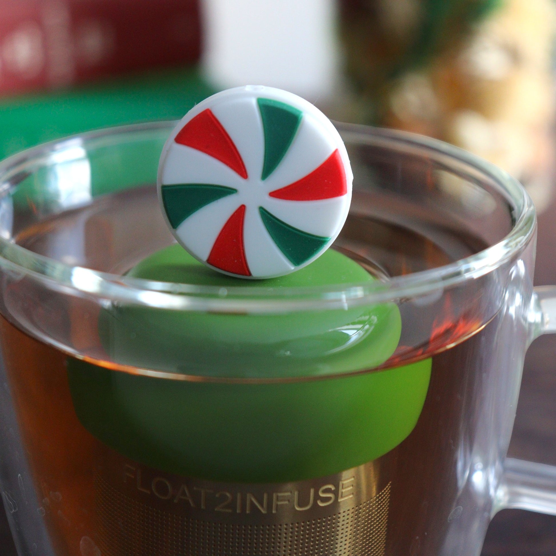red, white, and green striped peppermint candy tea infuser for loose leaf tea with a fine mesh stainless steel tea strainer 
that reads FLOAT2INFUSE on a dark green base floating in a glass tea mug with books and Christmas decorations in the background