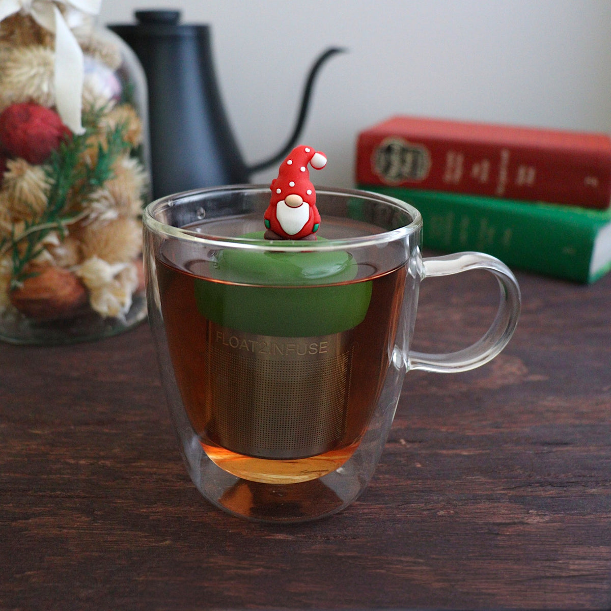 gnome with a long white beard wearing red with white polka dots on the hat and green mittens tea infuser for loose leaf tea with a fine mesh stainless steel tea strainer 
that reads FLOAT2INFUSE on a dark green base floating in a glass tea mug with books and Christmas decorations in the background