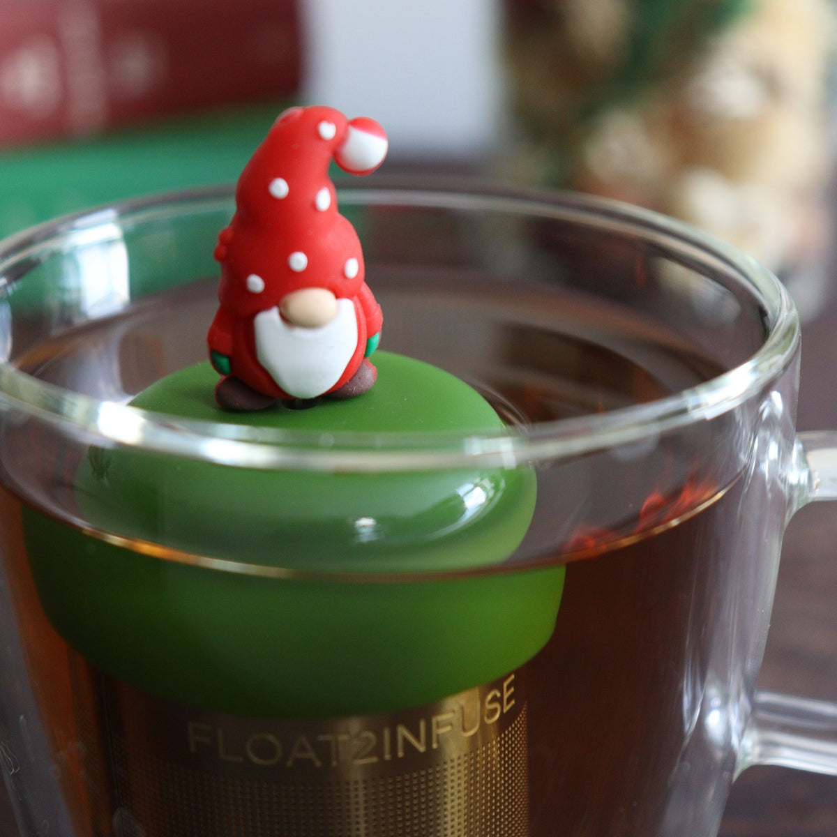 gnome with a long white beard wearing red with white polka dots on the hat and green mittens tea infuser for loose leaf tea with a fine mesh stainless steel tea strainer 
that reads FLOAT2INFUSE on a dark green base floating in a glass tea mug with books and Christmas decorations in the background