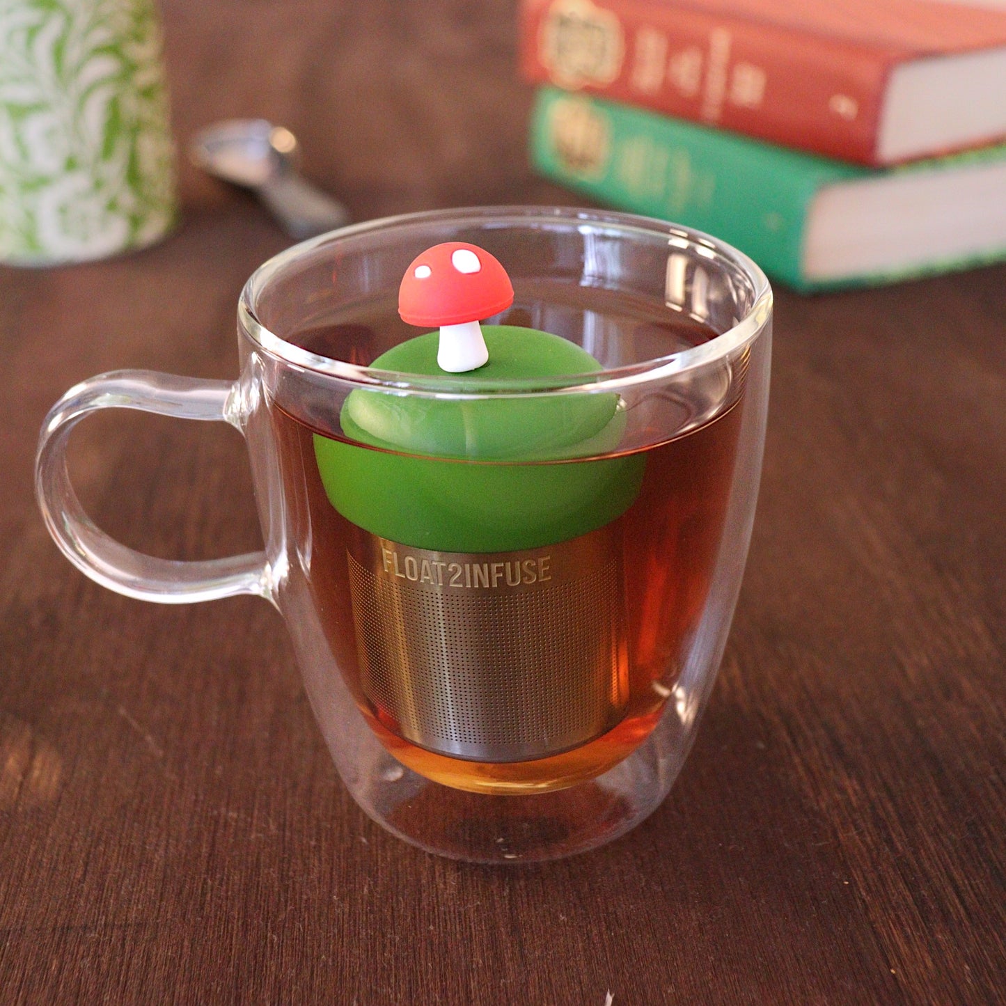 Red mushroom tea infuser for loose leaf tea with a fine mesh stainless steel tea strainer that reads FLOAT2INFUSE on a dark green base floating in a glass tea mug 
with a tea canister and books in the background
