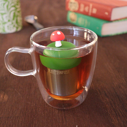Red mushroom tea infuser for loose leaf tea with a fine mesh stainless steel tea strainer that reads FLOAT2INFUSE on a dark green base floating in a glass tea mug 
with a tea canister and books in the background
