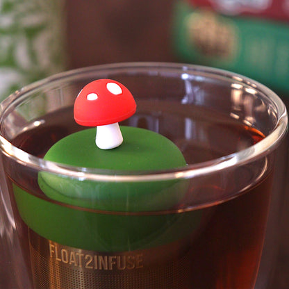 Red mushroom tea infuser for loose leaf tea with a fine mesh stainless steel tea strainer that reads FLOAT2INFUSE on a dark green base floating in a glass tea mug 
with a tea canister and books in the background