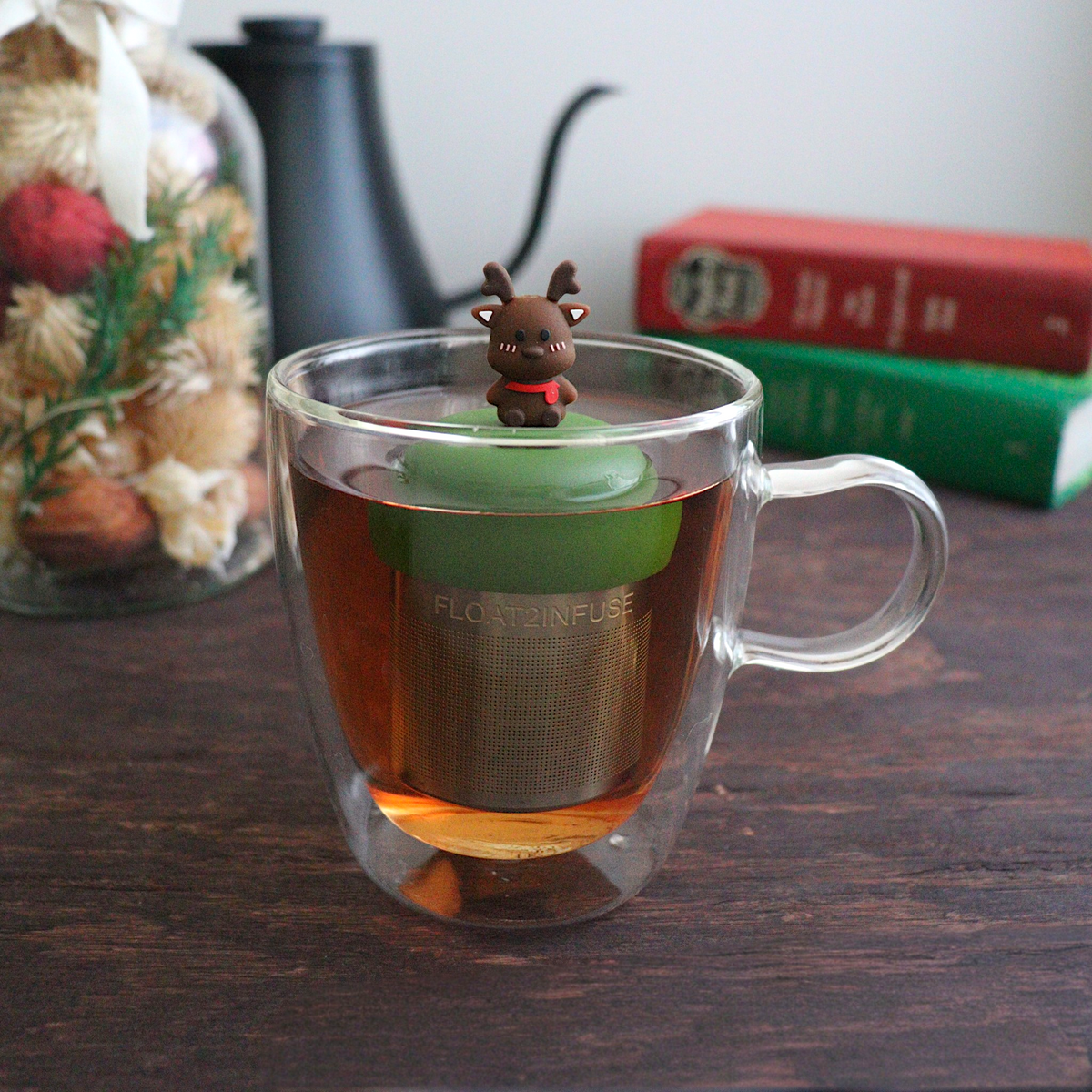 cute brown reindeer with a red scarf tea infuser for loose leaf tea with a fine mesh stainless steel tea strainer that reads FLOAT2INFUSE on a dark green base with books, a tea kettle, and christmas decorations in the background